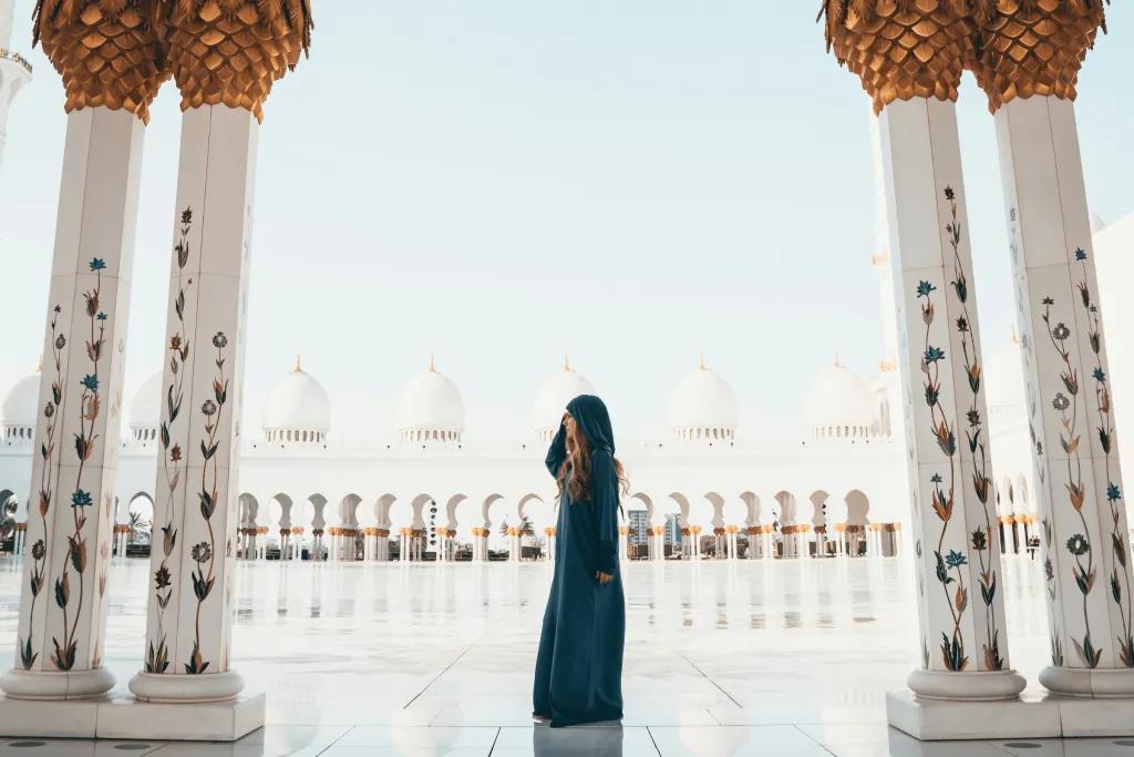Abu Dhabi Sightseeing There is a gorgeous sleek tiled floor which is so clean you can see a reflection of people in it. There's a woman wearing a robe which covers her whole body to be respectful to the culture. There are pillars which are white and gold with paintings of flowers growing up them and on the other side it is the same, with white domes on top of the pillars.