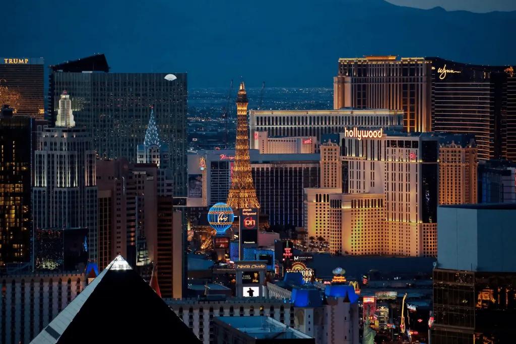 Las Vegas The strip of Las Vegas. There is a straight road with cars on it and a large pointy metal building coloured yellow with lighting, a replica of the Eiffel Tower. There is also a huge air balloon lighted in neon blue and yellow. There are many casinos and hotels as well.