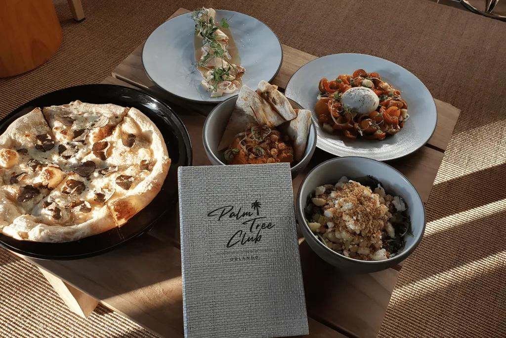 The Palm Tree Club Food There is a grey menu saying the Palm Tree Club on it and surrounding this there are 5 different dishes. One garlic bread, one mac and cheese and a few other delicious dishes.