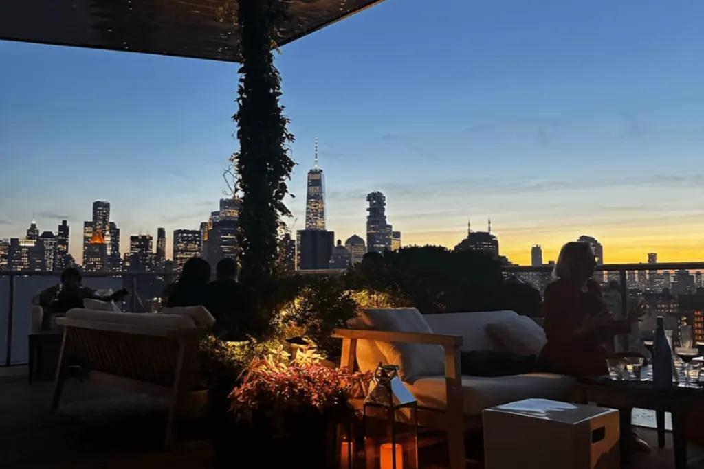 New York City Rooftop bar in new york city. The sky is a dark blue with a hint of yellow and orange as the sun sets in the evening. There are lamps on the ground with candles in them, flowers next to it and plants weaving around the wooden bar beams. This bar is outside and people are sat talking on sofas and the view shows the city skyline lit up.