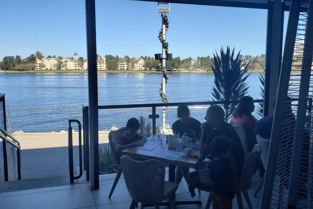 Summer House on the Lake Terrace A beautiful terrace with people dining at tables overlooking a large lake. There are posts with Christmas decorations swirling up them. On the other side of the lake there are pretty white buildings.