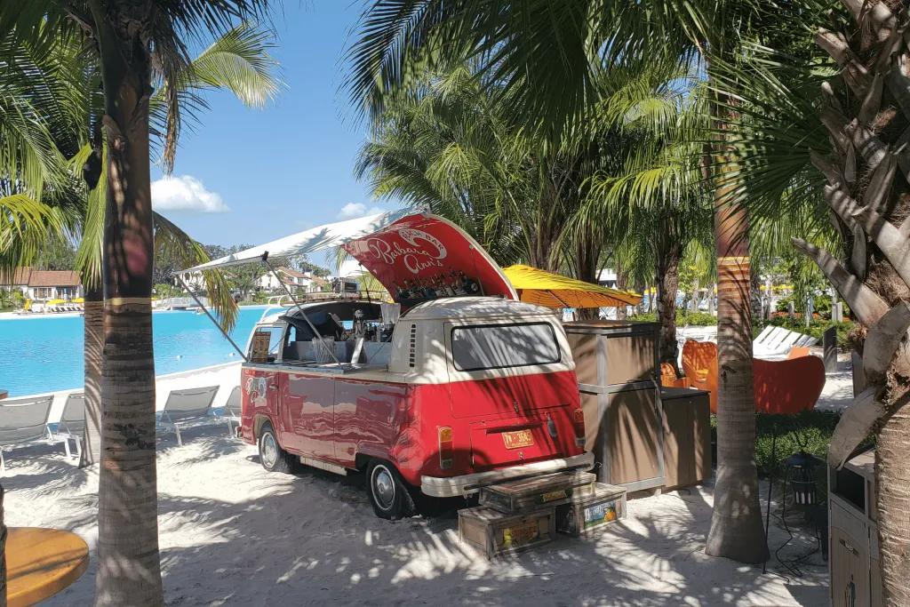 Evermore Resort Beach Bar On the white sand back there is a red campervan renovated into a bar. In the roof which is lifted up, there are bottles of alcohol. There are many palm trees surrounding this bar and the pool is in the background.