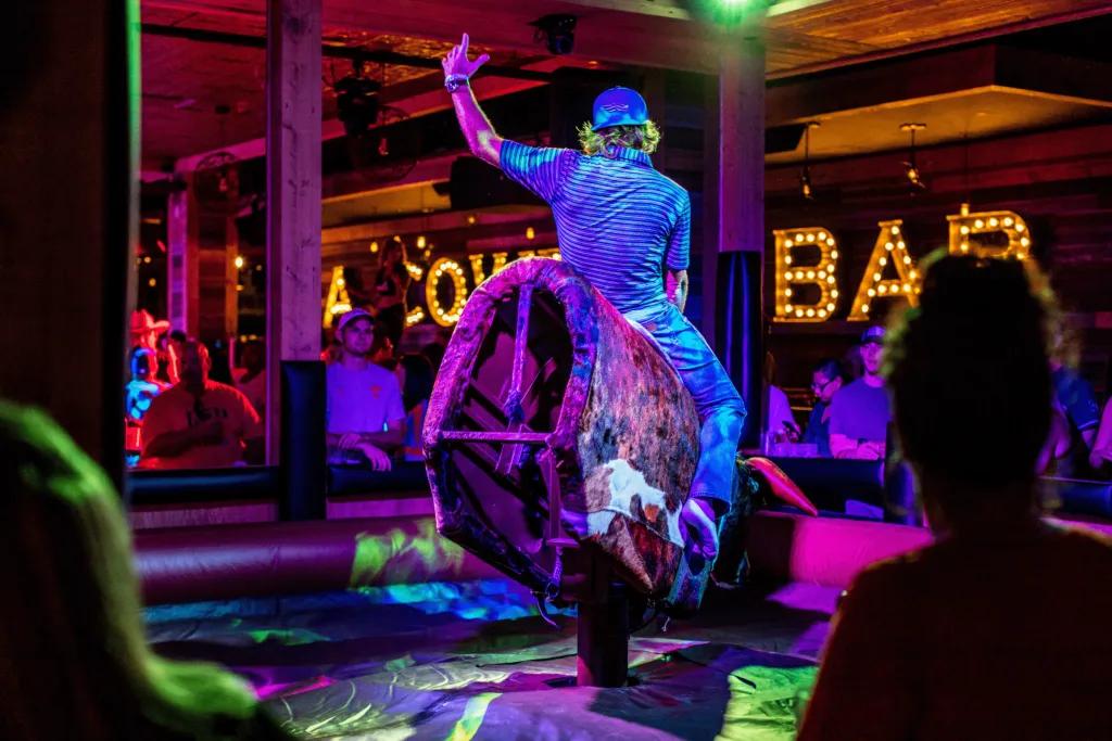 PBR Cowboy Bar Man with backwards cap on is on a mechanical fake bull, holding on tight and trying to stay on as it spins around and everyone is crowded watching around the sides of the black inflatable. There are beams everywhere and in the background you can see "A COWBOY BAR" in large lights on the wall.