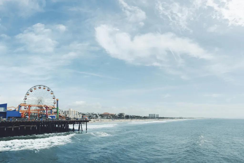 California There is the sea waves crashing onto a yellow sandy beach. There is also a long iconic pier with a funfair on it, including a Ferris wheel and small rollercoaster. In the far distance there are only a few large buildings..