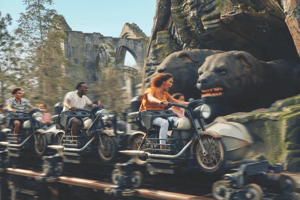 Hagrid's Magical Creatures Motorbike Adventure™ Guests on motorbike shaped vehicles and sidecars zooming past a three faced giant dogpoking out of a cave and there are ruins in the background.