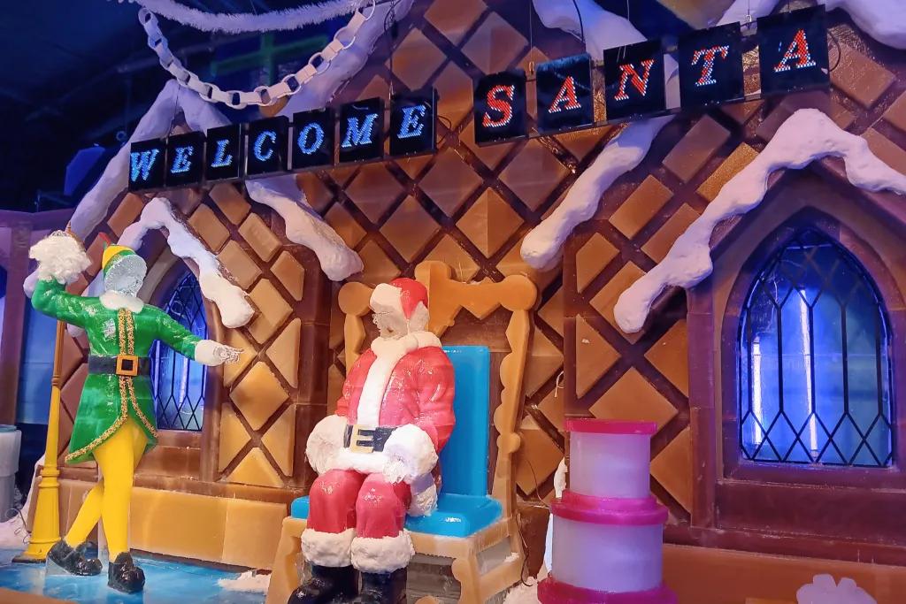 ICE! at Gaylord Palms Resort Ice sculpture of santa sat on a throne in front of a gingerbread house. Above the gingerbread house, there are blue and red signs saying "welcome Santa" and to the left, there is a buddy the elf sculpture throwing a lump of snow at Santa.