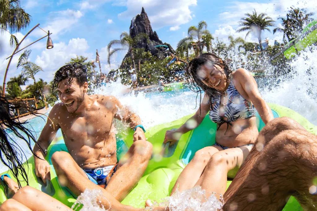 Volcano Bay 4 people sat in a big rubber ring heading into a large pool of water which is splashing them as they cling on. In the background there is a large brown/grey coloured volcano.