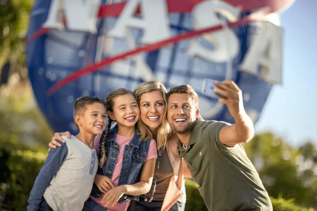 Kennedy Space Center Family of four smiling brightly, 2 kids and 2 parents taking a selfie in front of a large blue globe statue with red lines zooming across and the word NASA.