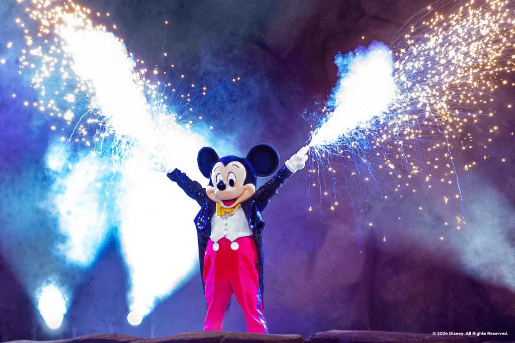 Mickey Mouse in Fantasmic! Mouse with black ears and beige face wearing a long black coat, red trousers and a white shirt. He has sparklers coming out of both of his hands.