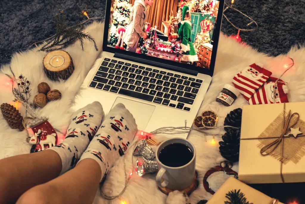 Christmas Movies Someone sat on a white blanket with christmas lights, watching a christmas movie on their laptop with decorations around like stripey socks and presents