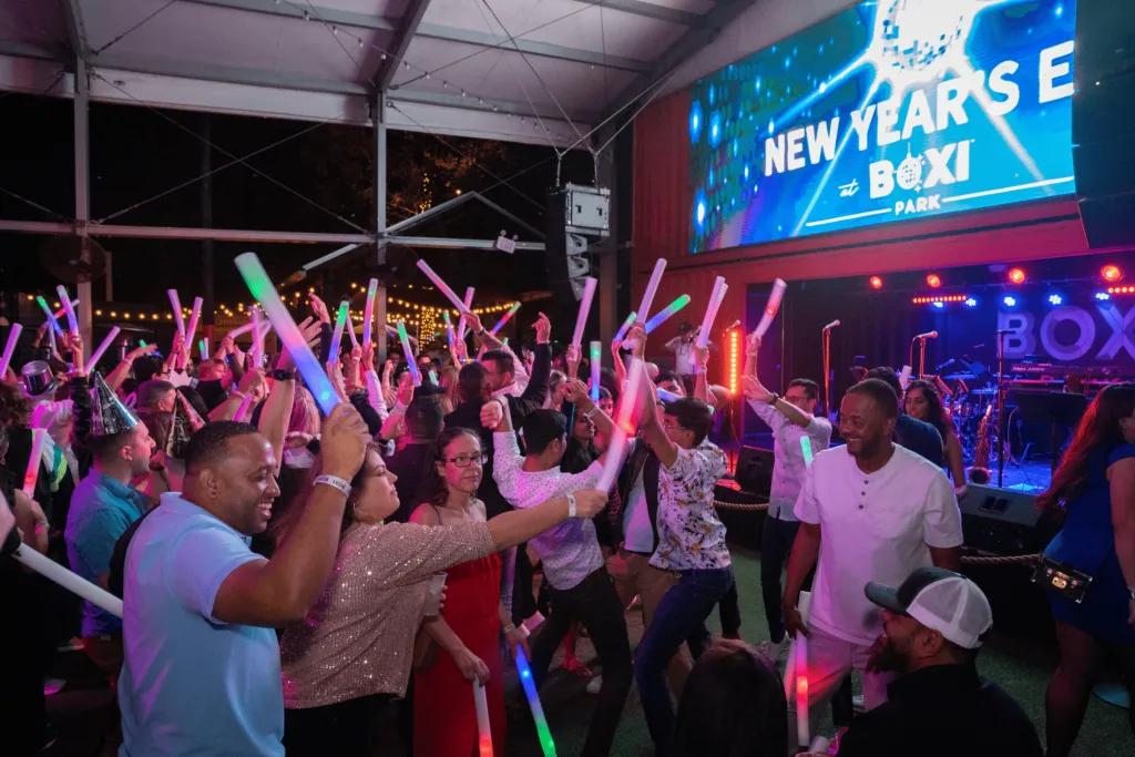 New Year’s Eve at Boxi Park Crowd of people holding up light sticks and dancing looking towards a screen which says New Years Eve Boxi and a sparkling disco ball.