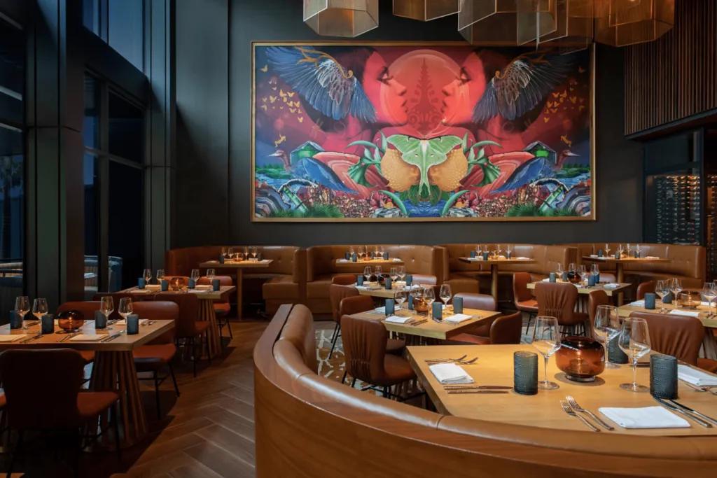 Baçan A large restaurant with huge painting on the wall which includes two red people symmetrically looking at one another. The tables are light wood and dark chairs.