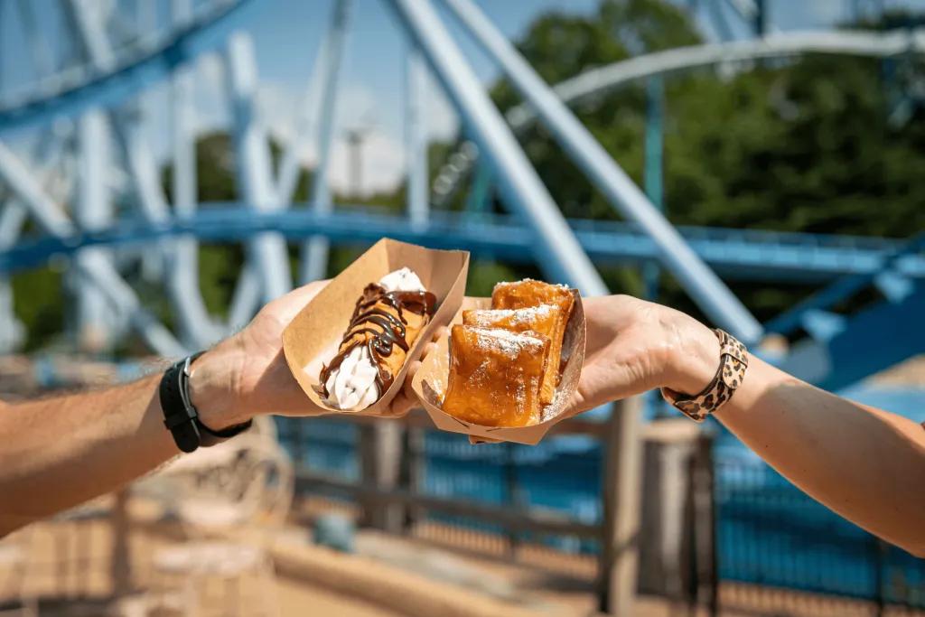SeaWorld Seven Seas Food Festival Two hands holding two dishes in front of a rollercoaster track. One is beignets and the other is a crepe with whipped cream on.