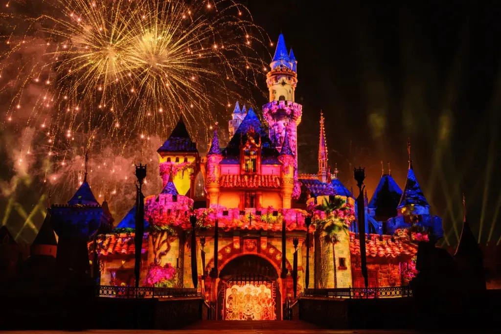 Wondrous Journeys Show Das kleine rosafarbene Schloss mit den blauen Dächern wird mit einer Projektion von Encanto beleuchtet und in der Mitte steht die Familie Madrigal in einem Familienporträt zusammen. Dahinter füllt ein gelbes Feuerwerk den Himmel.