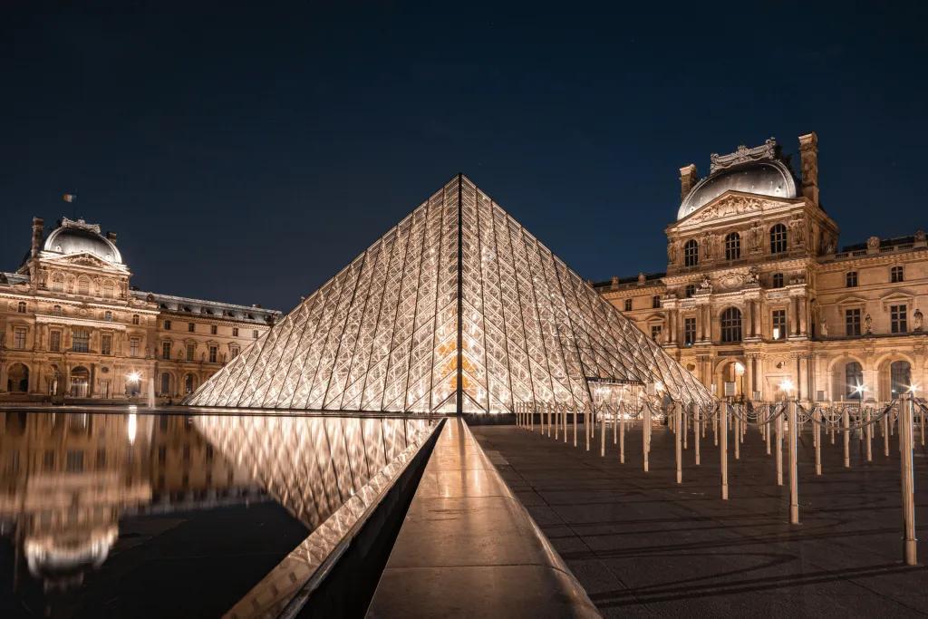 Louvre Museum A glass panelled pyramid building in the centre of old fashioned castle like buildings. It is reflecting in the small pond.