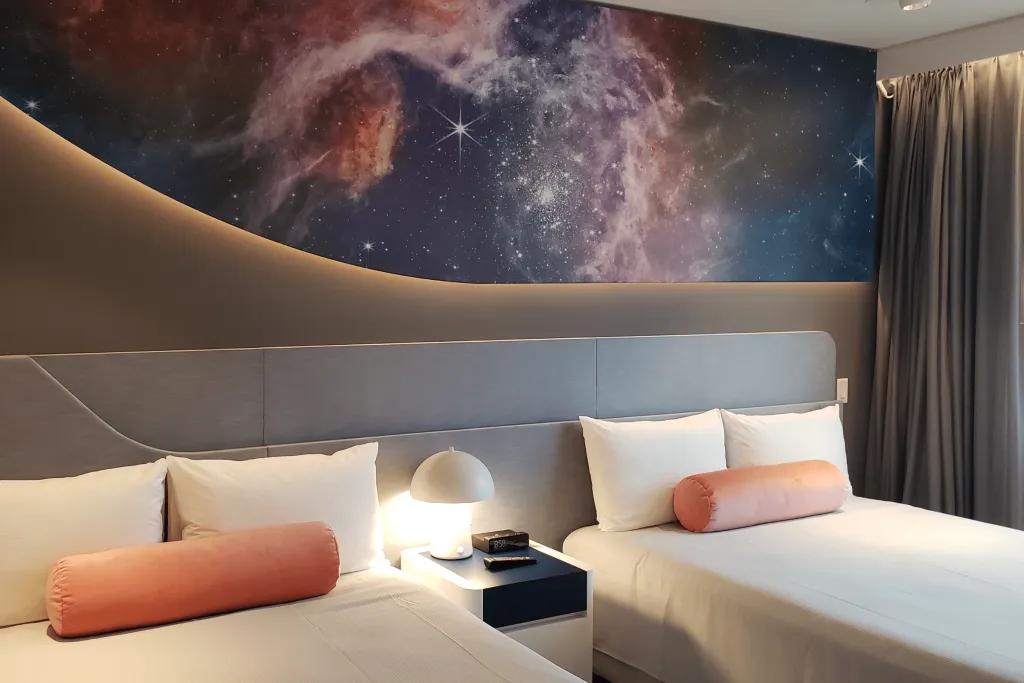 Stella Nova Resort Room with two white beds and orange cushions. There is a small white bedside table with a lamp and above on the dashboard there's images of stars in the universe.