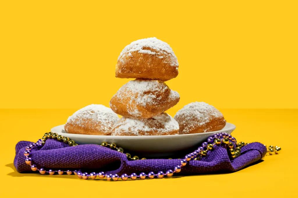 Mardi Gras Beignets A stack of beignets on a plate on top of a purple napkin and Mardi Gras beads