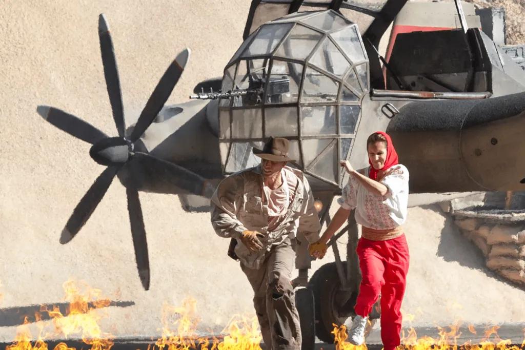 Indiana Jones Epic Stunt Spectacular Indiana Jones, a man dressed in explorer clothes and his sidekick, a woman wearing old fashioned clothes, running from a burning aeroplane about to explode, holding each others hands.