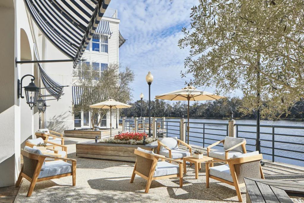 The Inn at Celebration Outside the large white hotel on the edge of a large lake in the sun, there are clean cream decking chairs, tables and umbrellas.