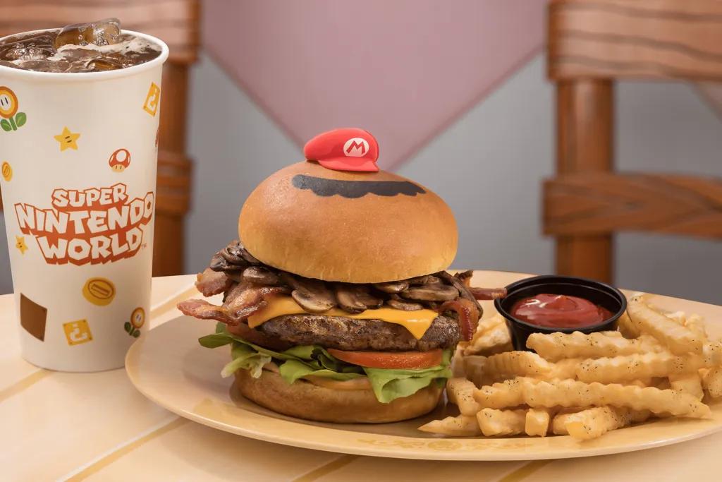 Mario Burger A large burger with: a brioche bun, large beef patty, cheese, mushrooms, lettuce, tomato and a little mario red hat on top. There are fries and a small black pot of ketchup. There is also a large cup of coke.