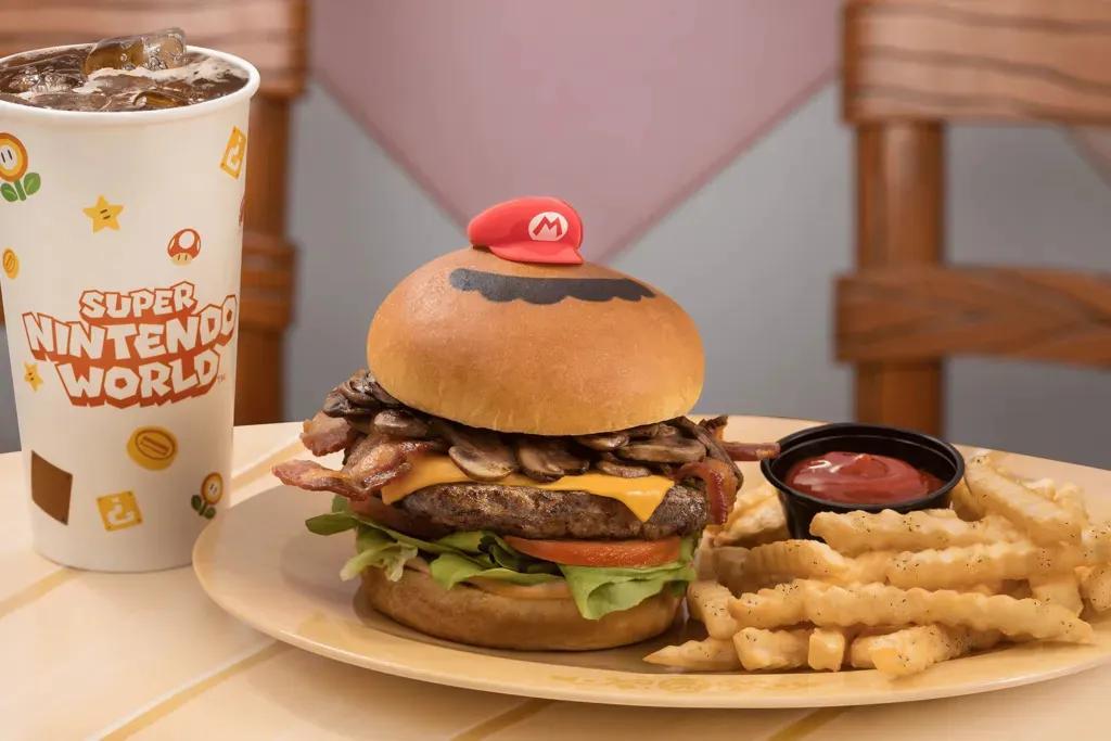 Mario Burger  Ein großer Burger mit einem Brioche-Brötchen, einem großen Rindpatty, Käse, Champignons, Salat, Tomate und einer kleinen roten Mario-Mütze darauf. Dazu gibt es Pommes und einen kleinen schwarzen Topf mit Ketchup. Außerdem gibt es einen großen Becher Cola.