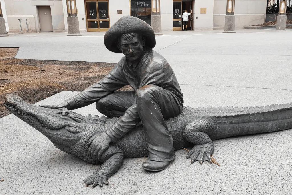 Bunk Baxter Statue Statue of man sat on alligator holding its head and arm, wearing a large hat.