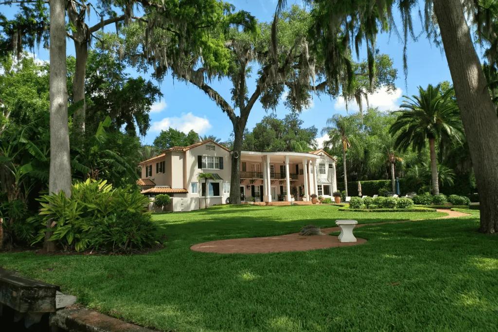 Winter Garden Scenic Boat Tour A large cream mansion with a lot of garden space, with trees and small white benches.