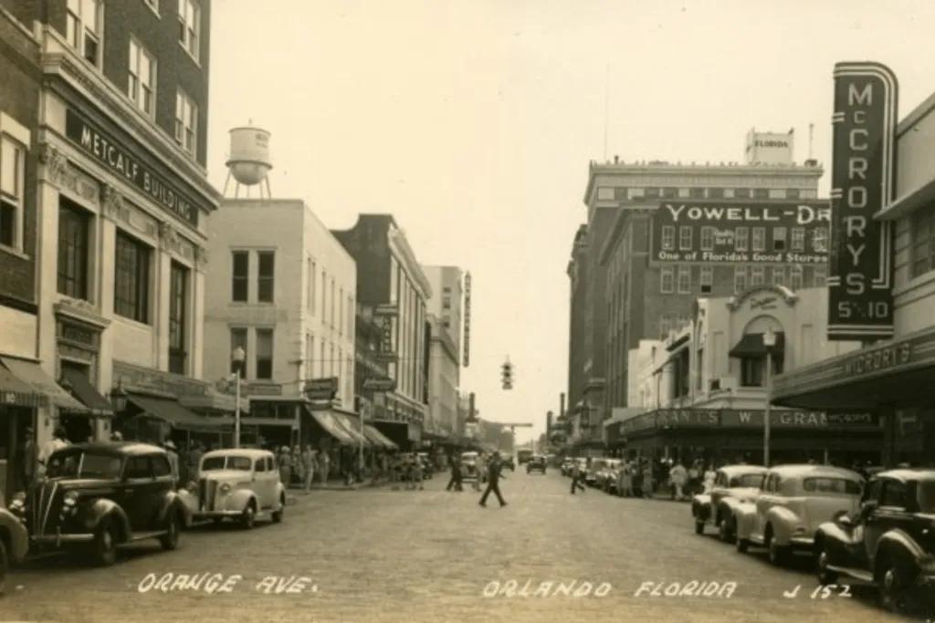 Orlando in the 1920's A black and white image of a street with pedestrians crossing in front of old fashioned beetle cars. There is old fashioned buildings, one with a water tower on top of it.