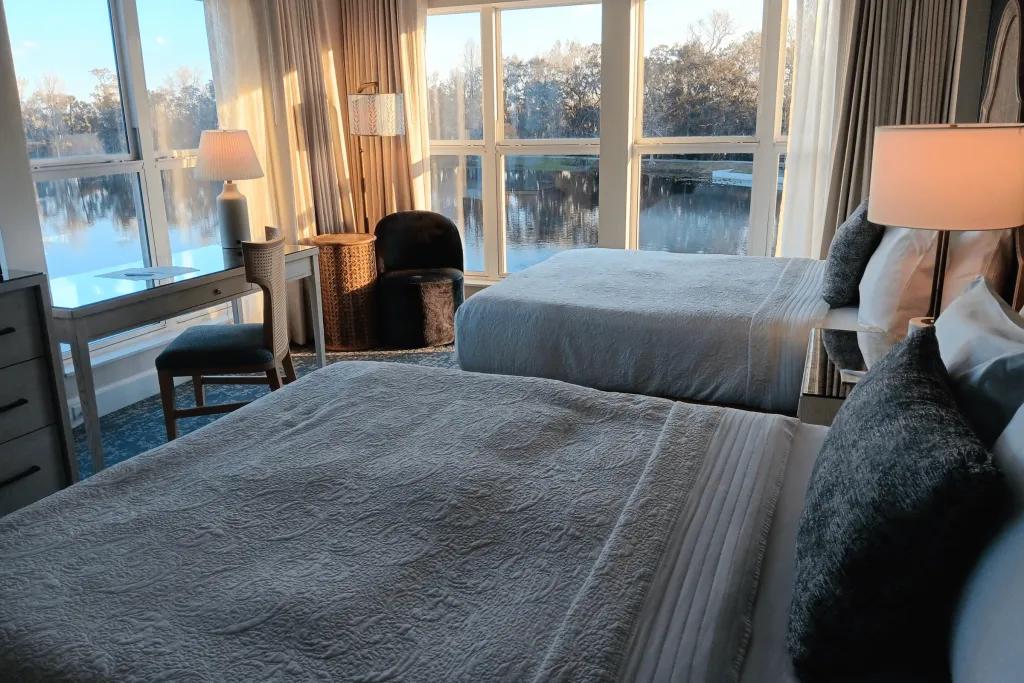 The Inn at Celebration Rooms Two beds in a hotel room with patterned sheets and green pillows on facing out towards to 2 large windows from floor to ceiling overlooking the beautiful lake.