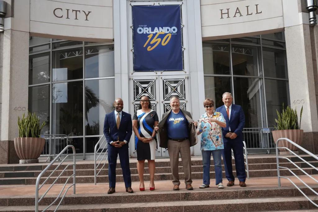 Mayor Dyer leads start of 150th anniversary at City Hall Mayor stood in a shirt celebrating the 150 year anniversary of ORLANDO with four other people. The City hall is behind decorated in banners saying Orlando 150.