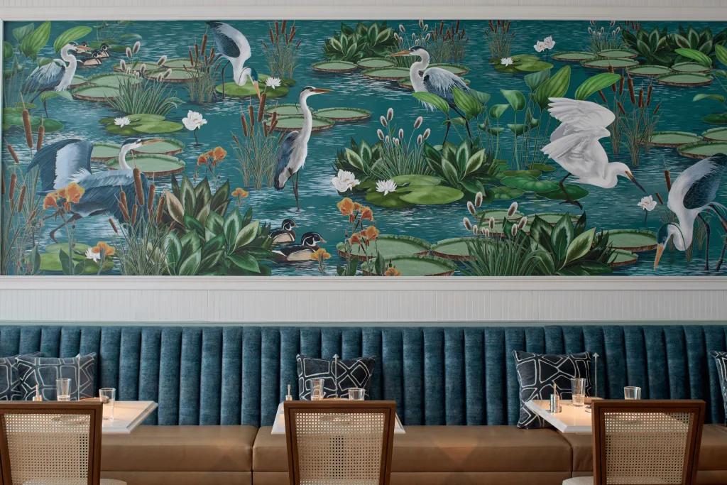 The Inn at Celebration Restaurant A restaurant with small table decorated with glasses, there is a booth side and a chair for each small table. There's a large wall behind full of art of herons in a lake.