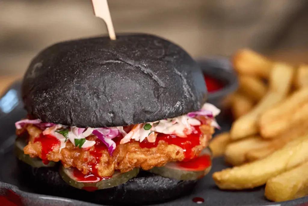 Blutorangen-Hähnchen-Sandwich Schwarzes Burgerbrötchen mit gebratenem Hähnchen, Krautsalat, Zwiebeln, Essiggurken und einer leuchtend roten Soße. Dazu gibt es Pommes und einen roten Dip.