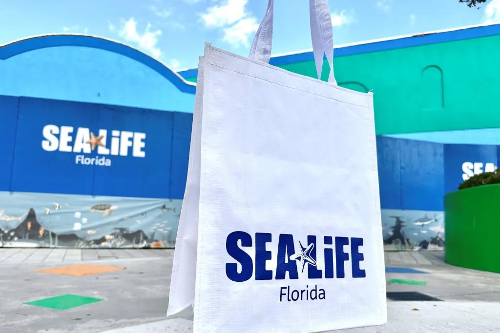 SEA LIFE Florida Huge blue building with SEA LIFE logo on it and a star fish. Under there is Florida label. There is a white tote bag being held up in front with a matching logo on it.