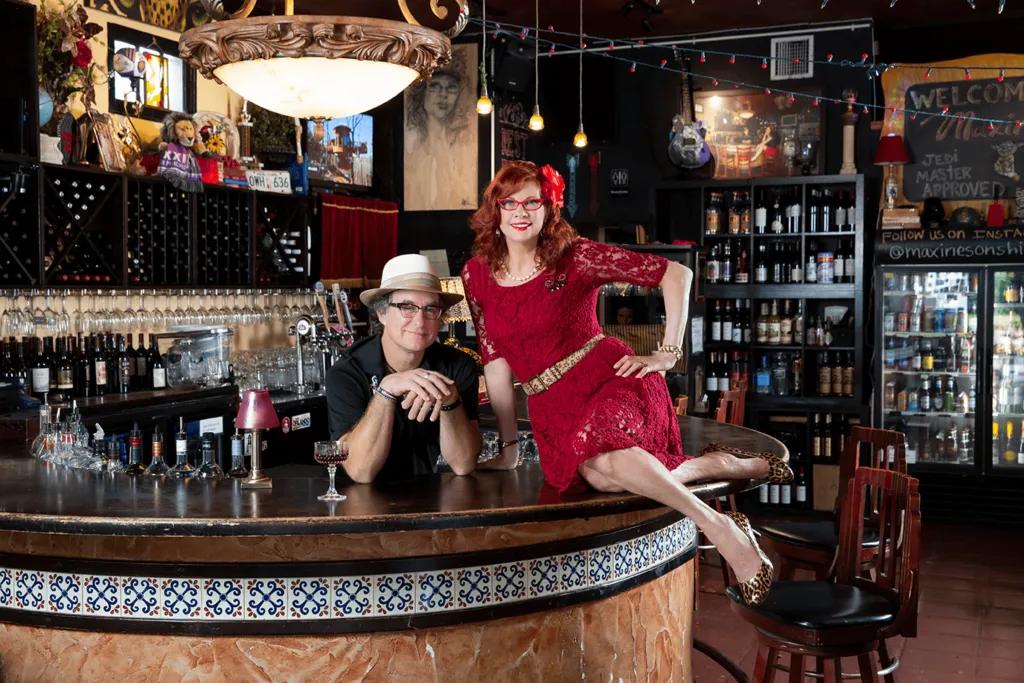 Maxine’s On Shine Woman in red dress with red flower in her hair sat on oval shaped bar, with bar stools behind her. A man is stood leaning on the bar from inside it.