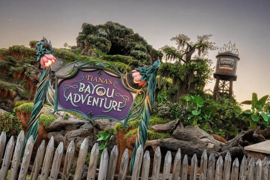Tiana's Bayou Adventure A sign made out of statue flowers which says "Tiana's Bayou Adventure". In the background there is a wooden water mill with a tiara on top. There is also a log flume ride covered in plants and flowers.