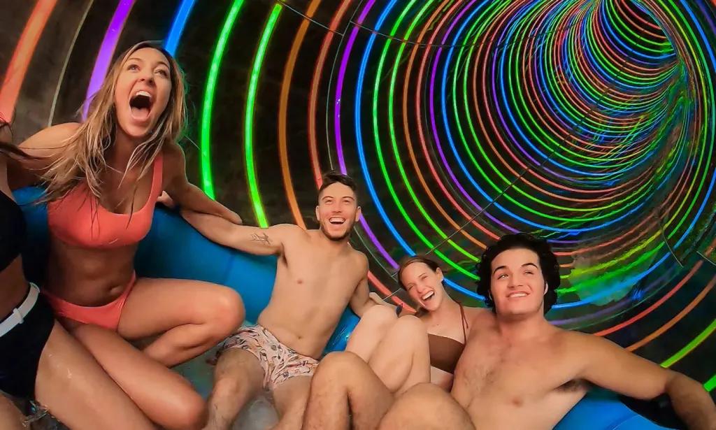 Profile Plunge People sat in a large round rubber ring facing one another in a slide tube that is rainbow coloured with neon colours.