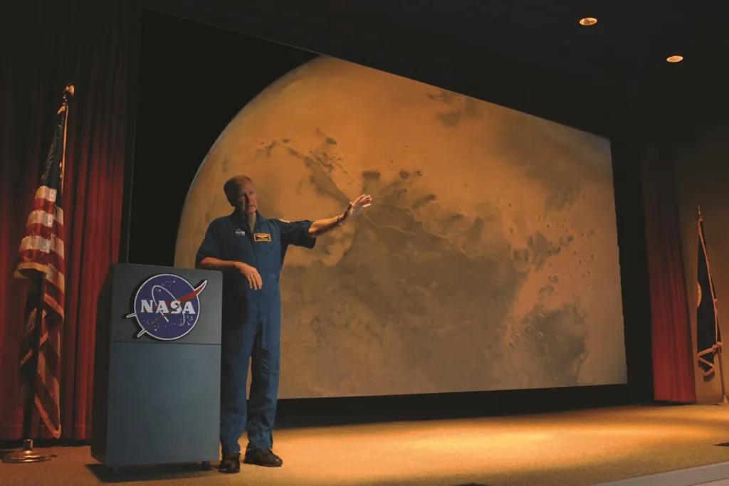 Astronaut Encounter Man stood in space suit on stage with planet presented on screen behind him.