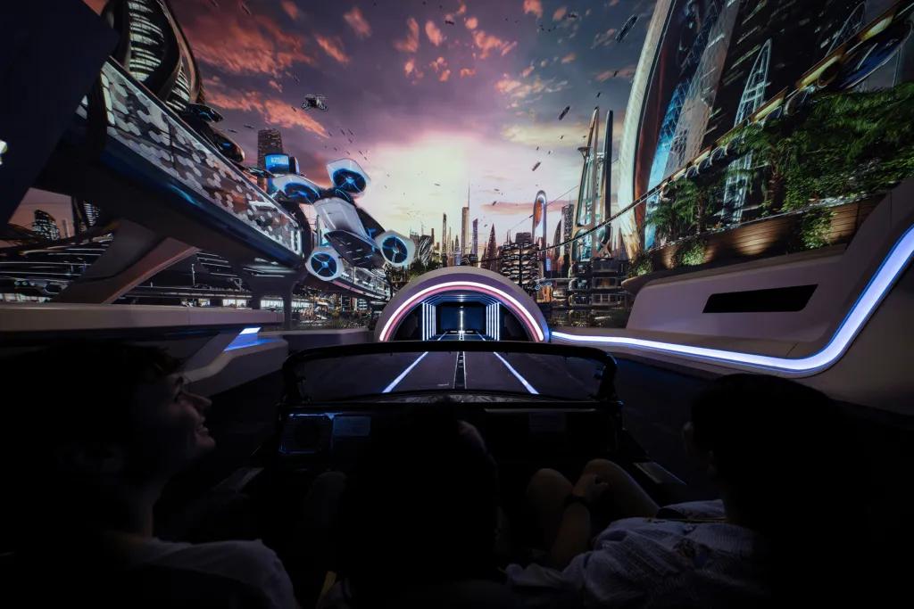 Test Track Update People sat in a car shaped vehicle, 4 in a row, there is a large screen with a modern, futuristic world on it and a small tunnel where the track leads to.
