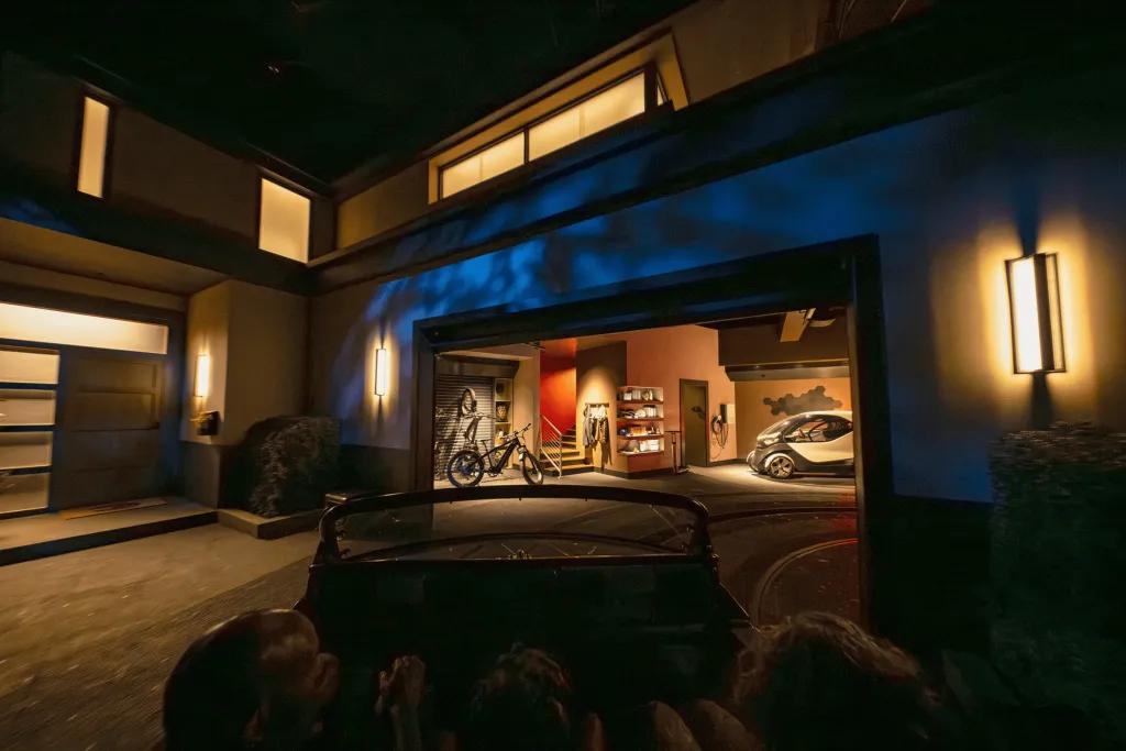 Test Track Updated People sat in car shaped rollercoaster vehicle heading into a room where there are bikes and other things stored.