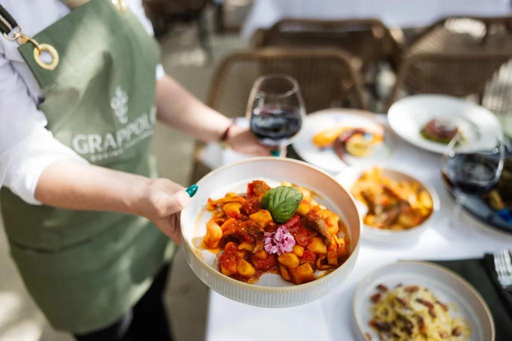 Grappolo Cucina Italiana A waiter holding a plate of pasta and a glass of wine