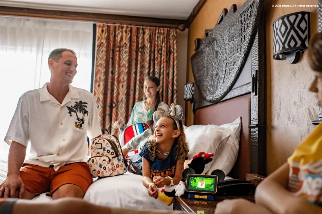 A smiling family relaxing in their Walt Disney World Resort hotel room, featuring a girl with Minnie Mouse ears sitting on the bed with a Mickey Mouse plush, showcasing the importance of rest for young families.