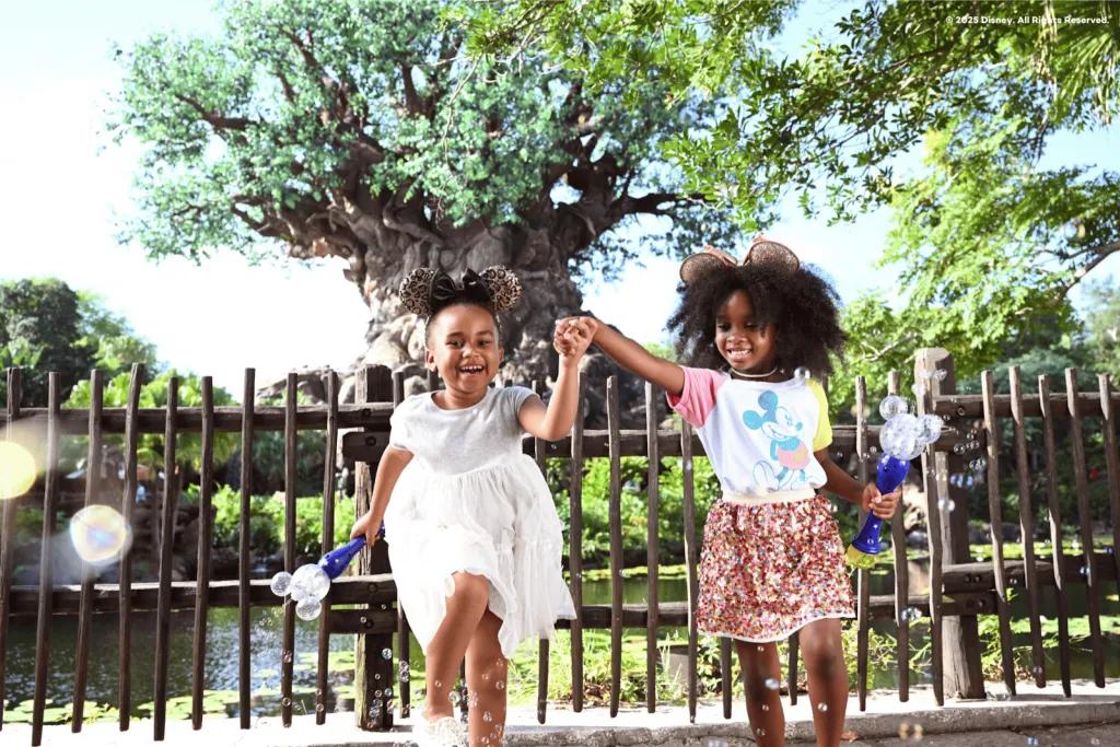 Two joyful young girls wearing Minnie Mouse ears and colorful park outfits, holding hands while playing with bubble wands in front of the Tree of Life at Disney's Animal Kingdom.