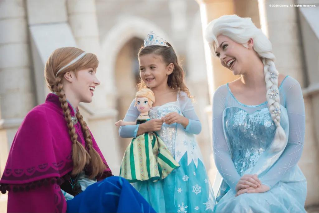 A young girl wearing an Elsa dress and a tiara, holding an Anna doll, smiling between costumed Anna and Elsa characters at Walt Disney World