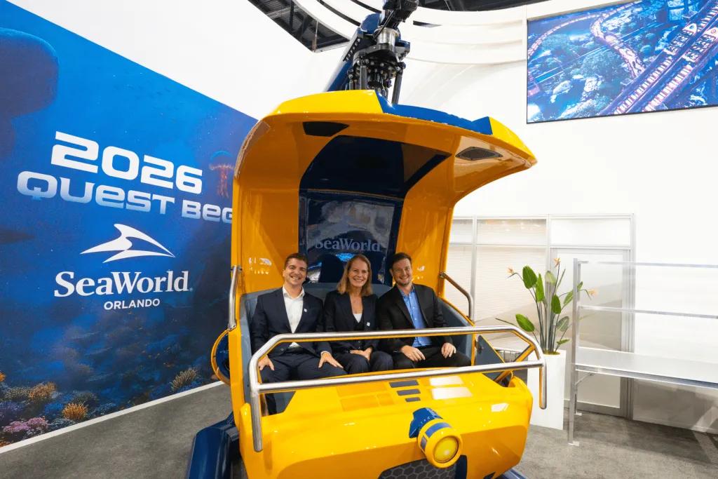 Yellow hanging ride vehicle with three grown adults sat on it and a screen behind with 2026 written on it. 