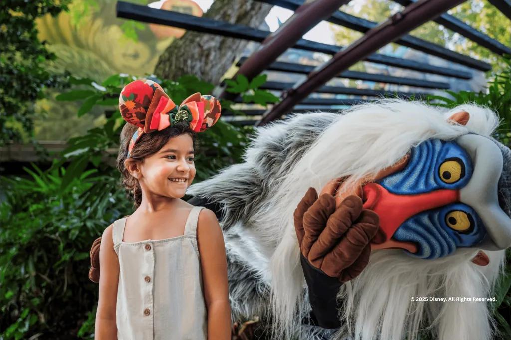 A smiling young girl wearing Minnie Mouse ears meeting the costumed character Rafiki from The Lion King at Disney's Animal Kingdom park.