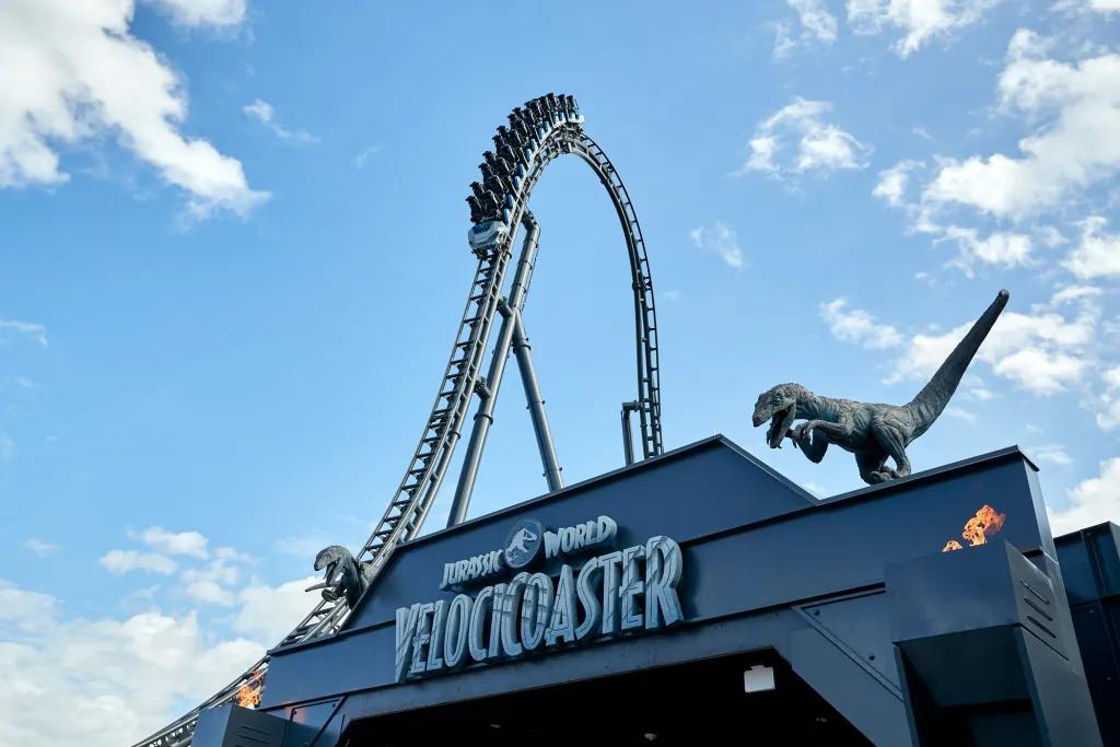 Rollercoaster diving above a steep drop with dinosaur statues on it and a sign saying "Jurassic World VelociCoaster"