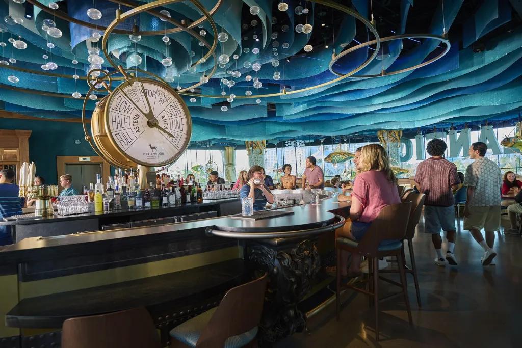 People sat in an underwater themed bar and restaurant with a large golden clock in the middle and fish hanging overhead.