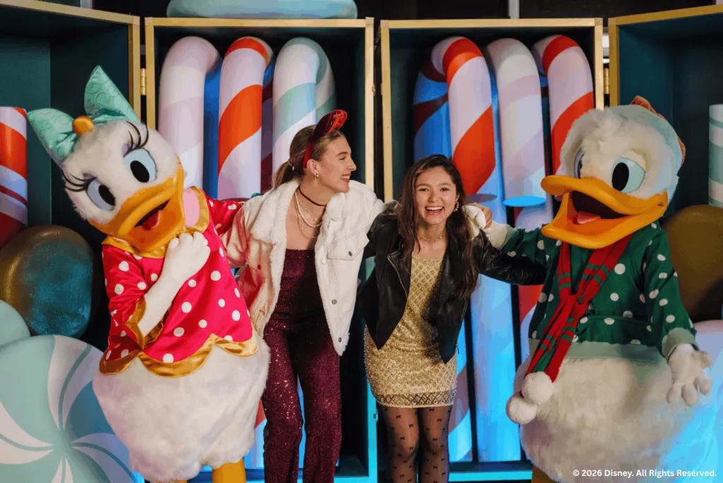 Two guests posing with Daisy and Donald Duck in holiday outfits against a candy cane backdrop