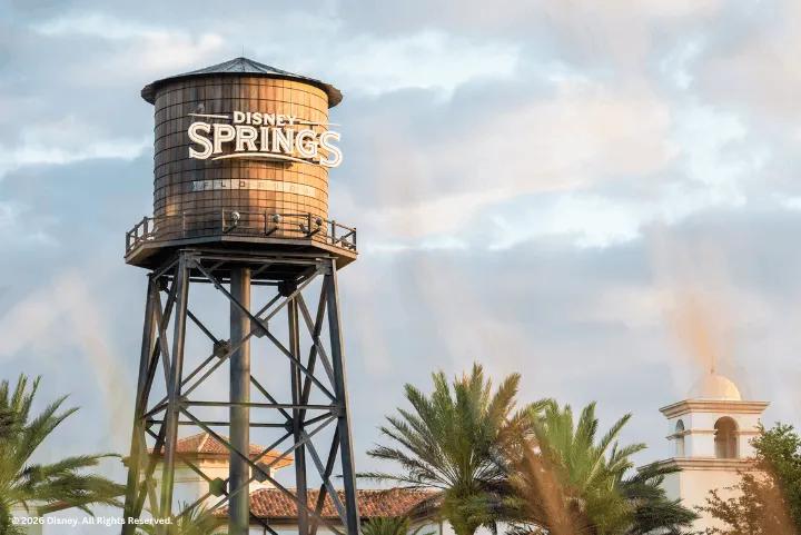 Disney Springs water tower sign against a cloudy sky, with palm trees and a building cupola in the foreground