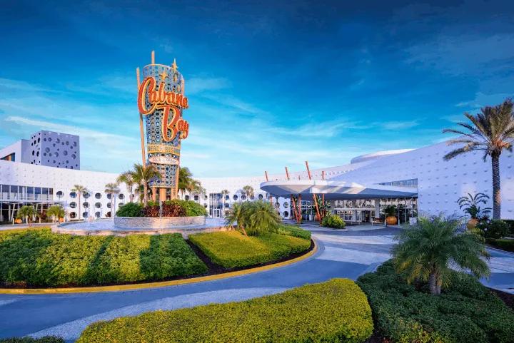 The front entrance of a retro-themed hotel featuring a tall, bright neon sign that reads "Cabana Bay Beach Resort," surrounded by a circular driveway, green landscaping, and white buildings with porthole windows under a blue sky